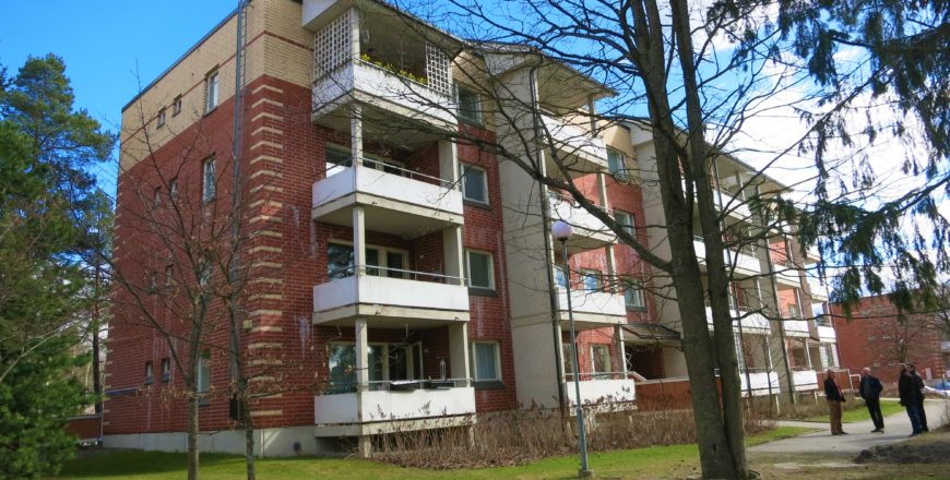 Krutkällarvägens flervåningshus i Esbo. Huset består av fyra våningar och är rött-gult-färgat. I varje våning finns vita balkonger. Gräsmattan och några stora träd finns på gården.