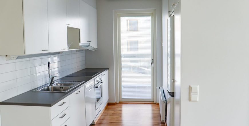 Köket i Koltgrändens lägenhet som är mestadels vitt utom den gråa bänkskivan. Genom dörren i köket går man till balkongen.