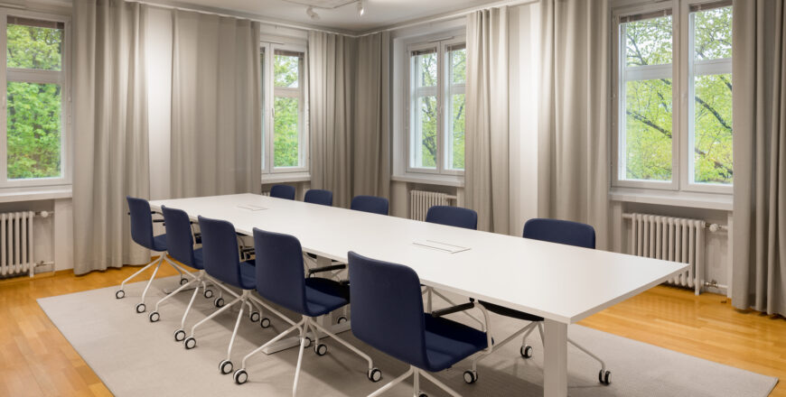 Pitkä kokoushuoneen pöytä, jonka ympärillä 10 tuolia. Tuoleissa on pyörät alla. Huone on kulmahuuone, jakahdella seinällä on yhteensä neljä ikkunaa kahteen eri suuntaan.