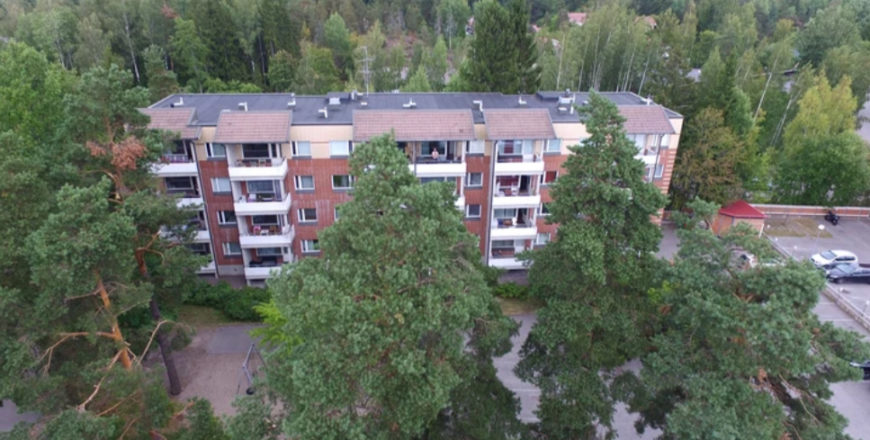 Flygfoto av Krutkällarvägens höghus som är omgiven av skogig landskap. Det finns torkningställ, lekplats och flera parkeringsplatser på gården.